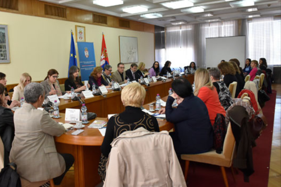 Veliki broj učesnika dijaloga sedi oko konferencijsog stola. U pozadini se vidi zastava Republike Srbije