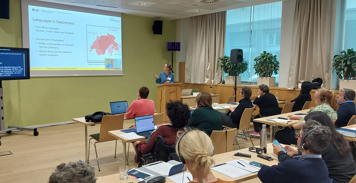 Predavanje na temu jezika u Švajcarskoj. Vidi se predavač i učesnici konferencije koji sede